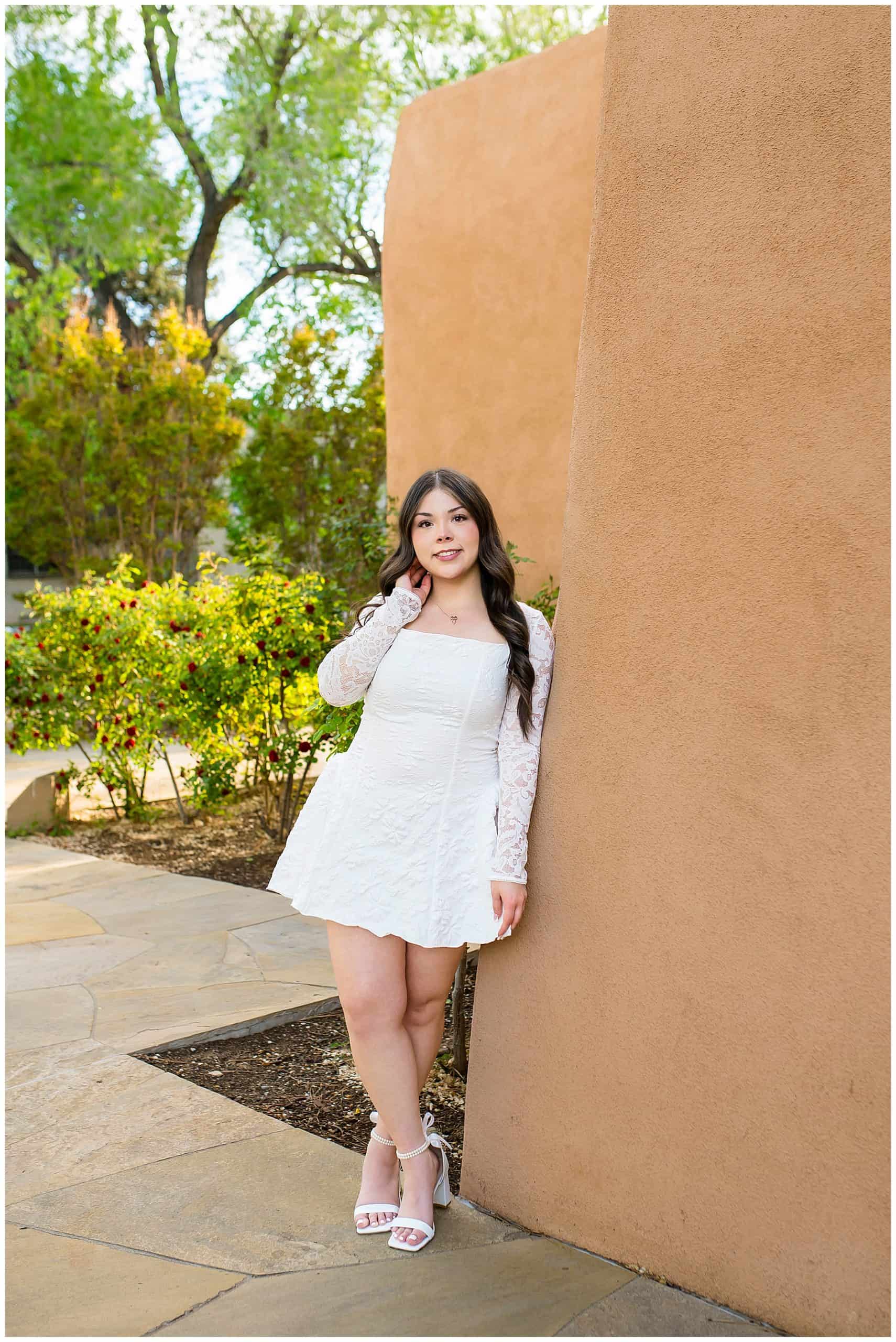 Senior girl in a white dress leaning against an adobe wall with vibrant red roses in Albuquerque.