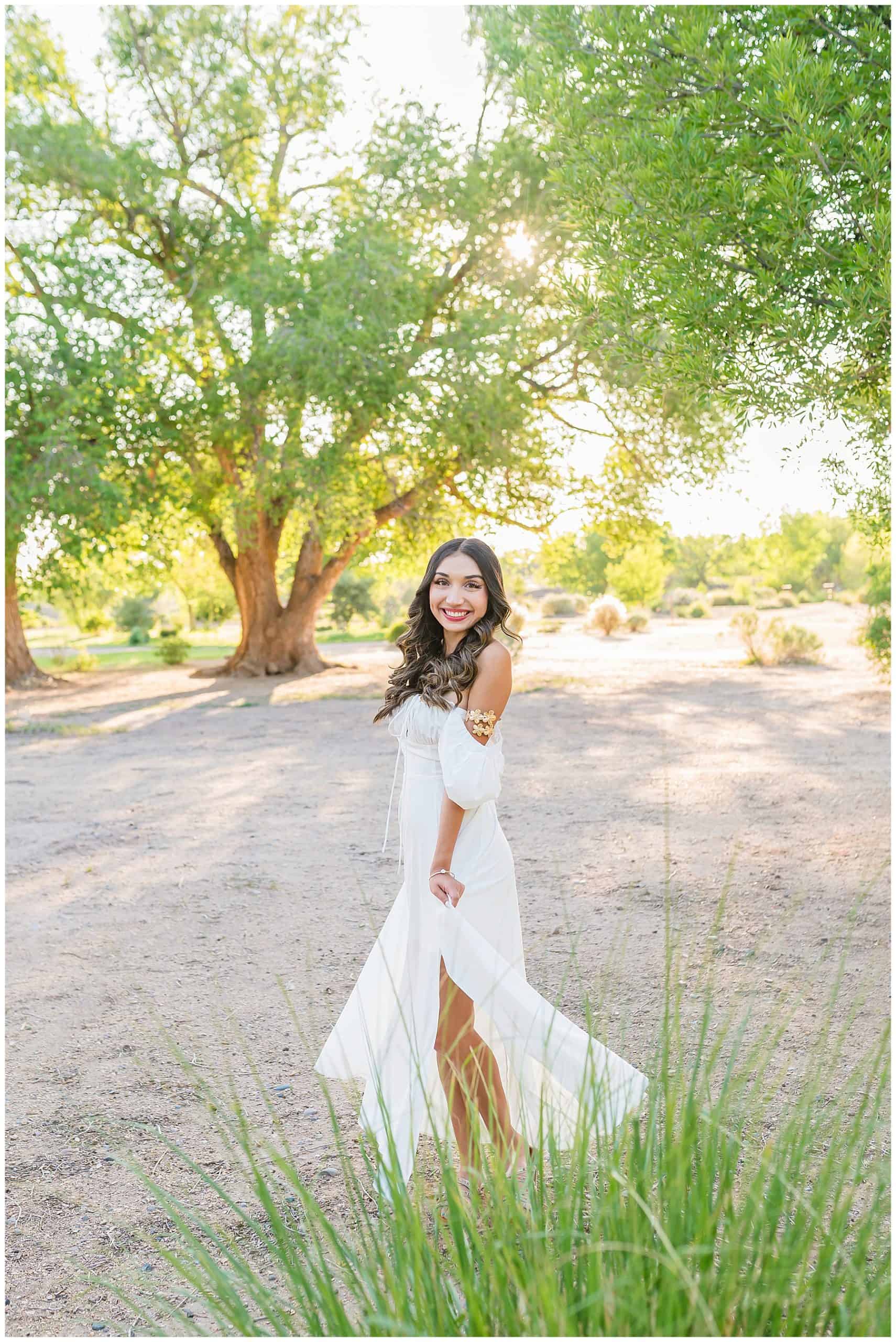 Senior girl wearing a white dress posing under a lush green tree at Bachechi Open Space during her Albuquerque senior session.